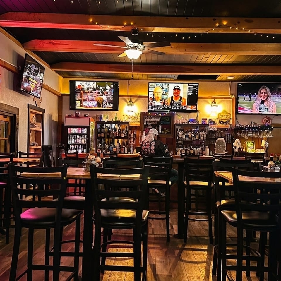 A dimly lit bar interior with high-top tables, chairs, multiple TVs, and a well-stocked bar.